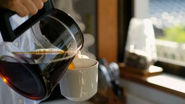 実写のカット。朝のキッチン。白いマグにコーヒーを注ぐ女性の手元。湯気が立つ。女性が窓辺でひと口飲んで「今日も、いい一日になりそう」とつぶやく。柔らかい朝の光。